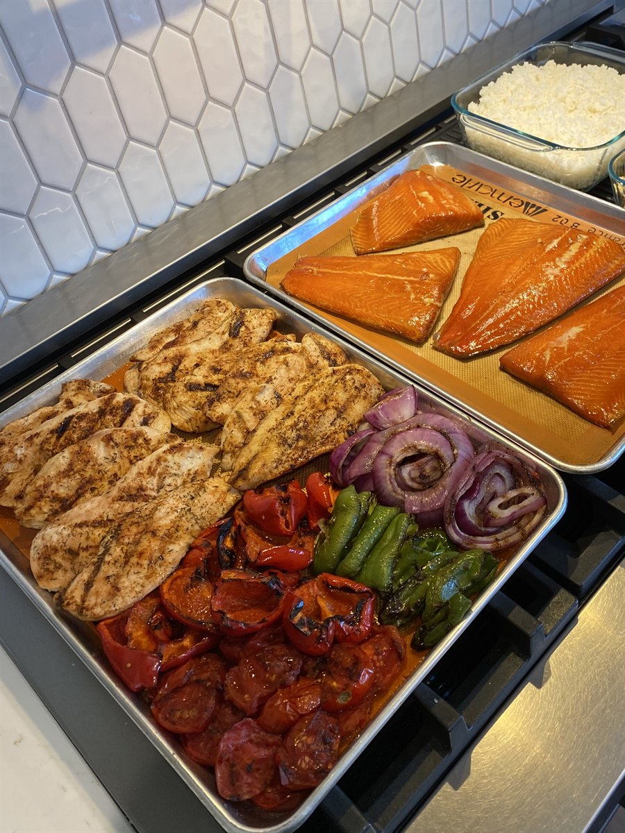 Two sheet pans on the stove: grilled chicken breasts with roasted peppers and red onion on one, salmon fillets on the other, with a dish of rice alongside